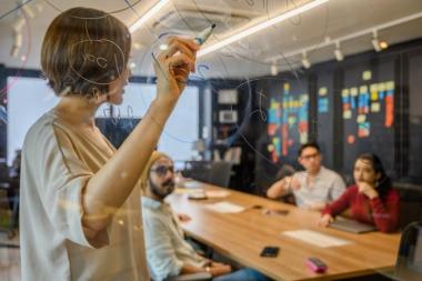 person writing on a glass whiteboard with colleagues in the background
