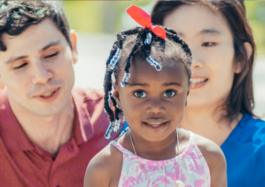 Little girl wearing a pink dress with a man and woman crouched behind her