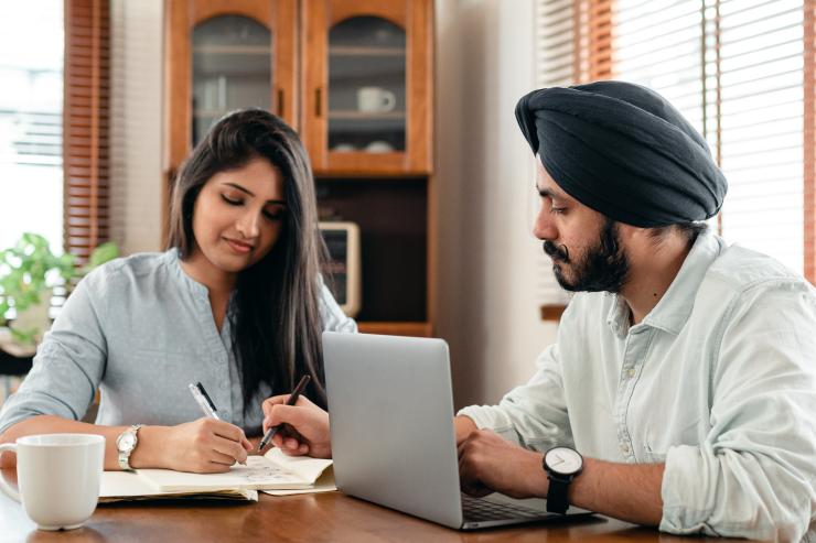 Man and woman working at a table. The woman is looking down towards the table whilst the man is looking towards her with a laptop in front of him, he's also wearing a headscarf.